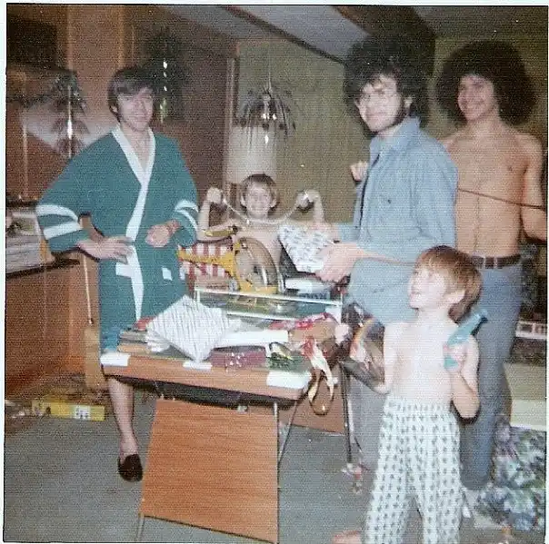 A grainy, vintage photo shows five people in a cluttered room, mostly shirtless or in loungewear, posing around a messy…
