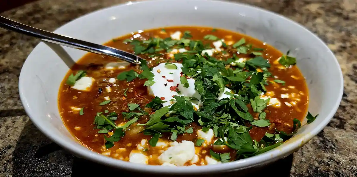 A close-up photo of a white bowl filled with a rich, reddish-brown soup, garnished with a dollop of white cream, chopped…