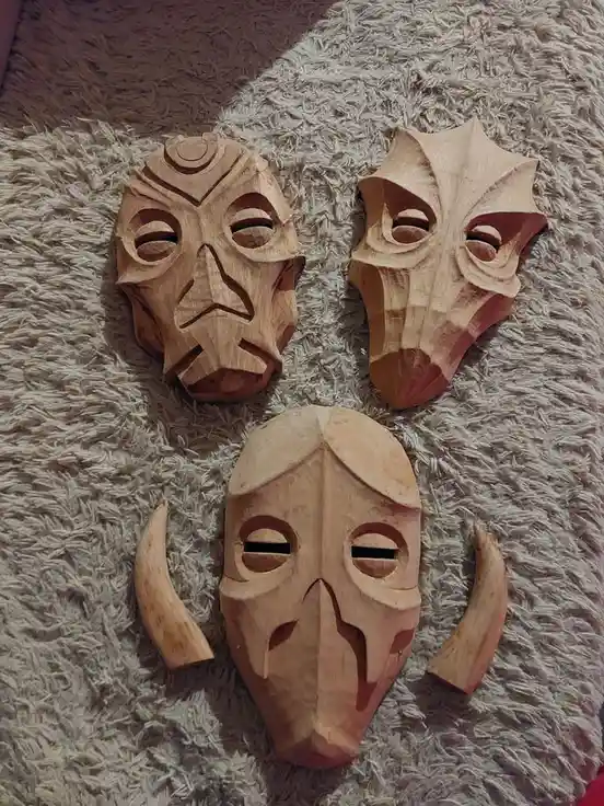 Three hand-carved wooden masks with stylized, angular features lie on a fluffy beige rug. The top-left mask has a…