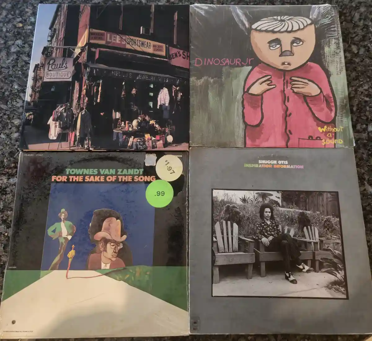 Four vintage vinyl record album covers are arranged in a grid on a speckled countertop. Top left shows a street scene with…