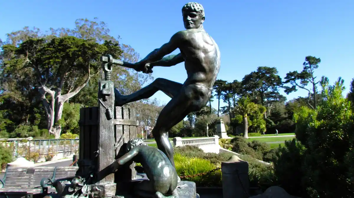 A bronze statue depicts a muscular man wielding a hammer against a wooden gate, while a smaller figure is pinned beneath it…