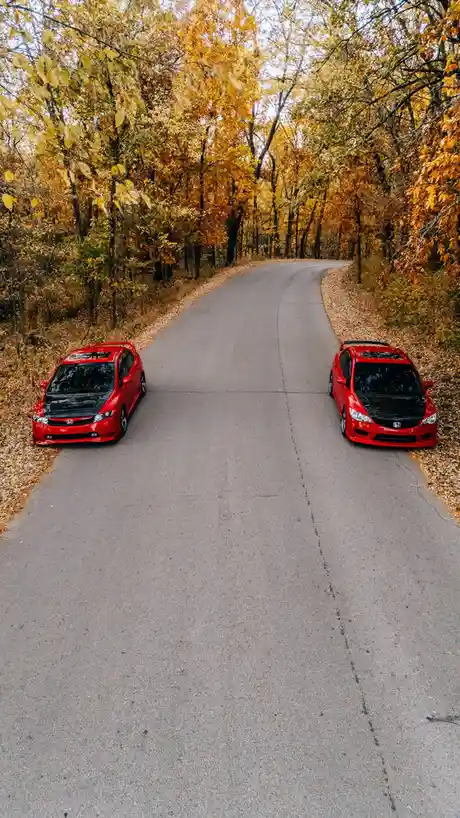 Two identical red Honda Civic Si cars are parked facing each other on opposite sides of a winding asphalt road surrounded by…