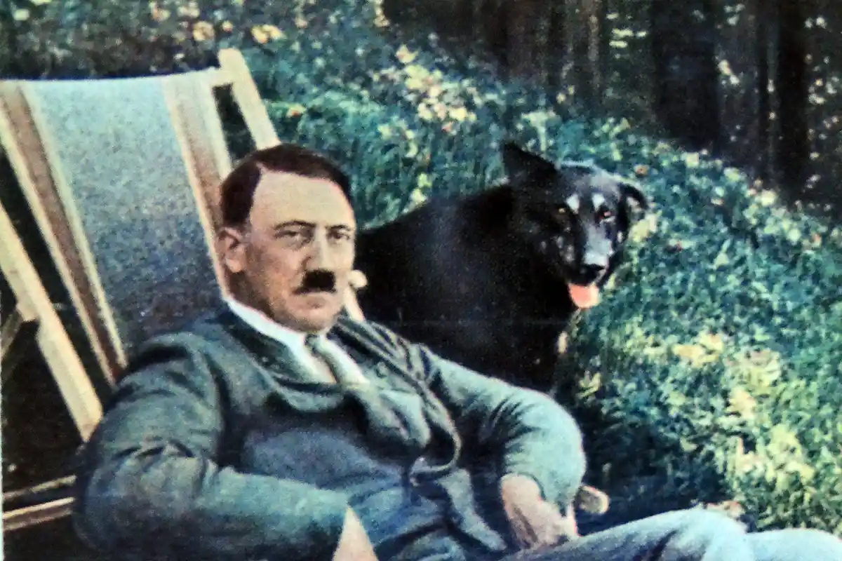 a historical photograph shows a man with a mustache sitting in a lawn chair outdoors, with a black dog standing behind him…