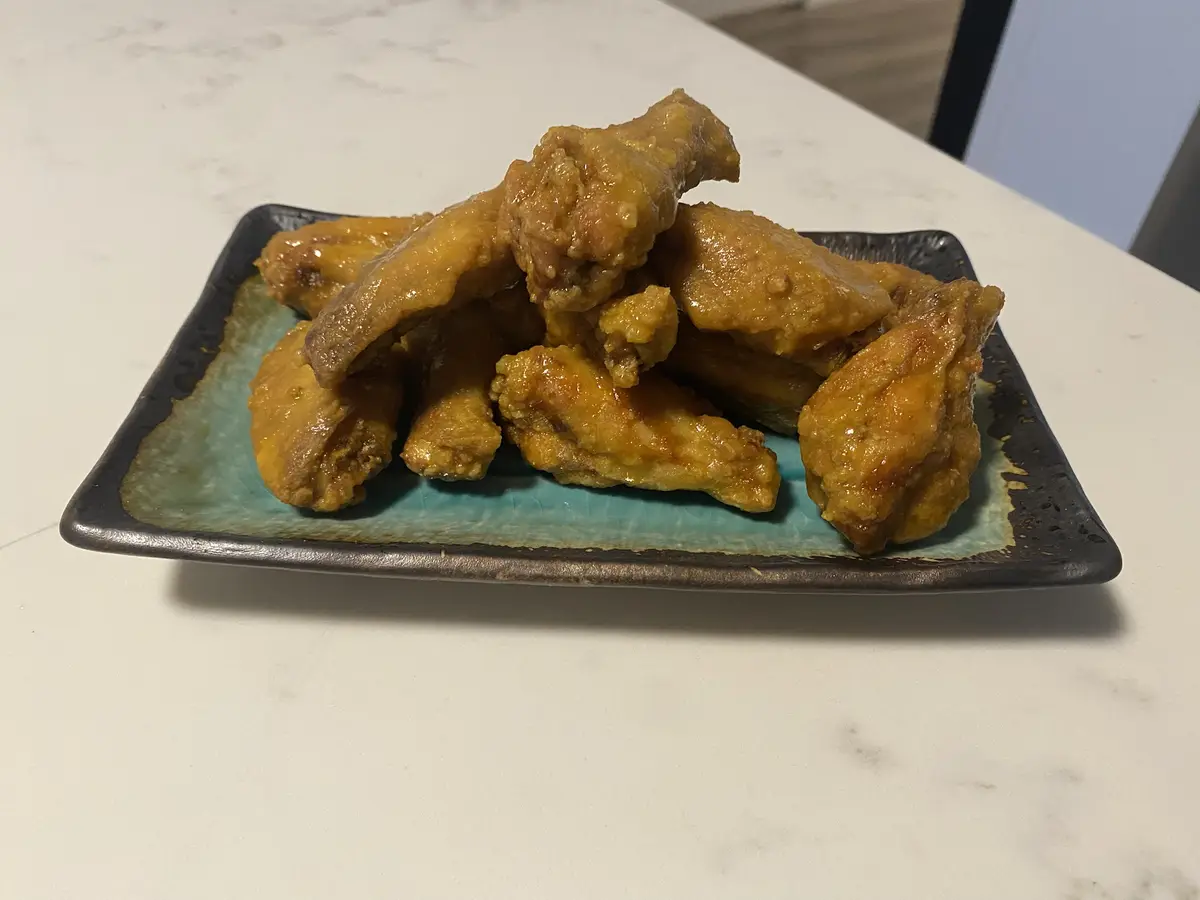 A plate of golden-brown, glossy fried chicken wings sits piled high on a rectangular ceramic dish with a teal center and…
