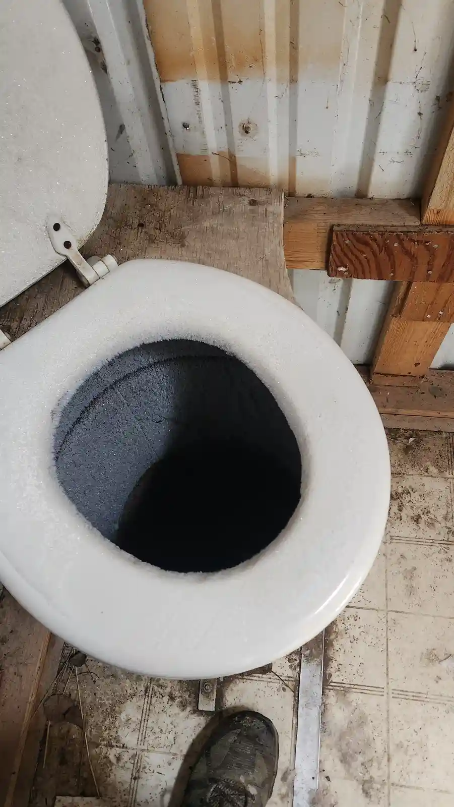 A close-up photo shows a white porcelain toilet bowl filled with a thick layer of ice, with the ice forming a dark, hollow…