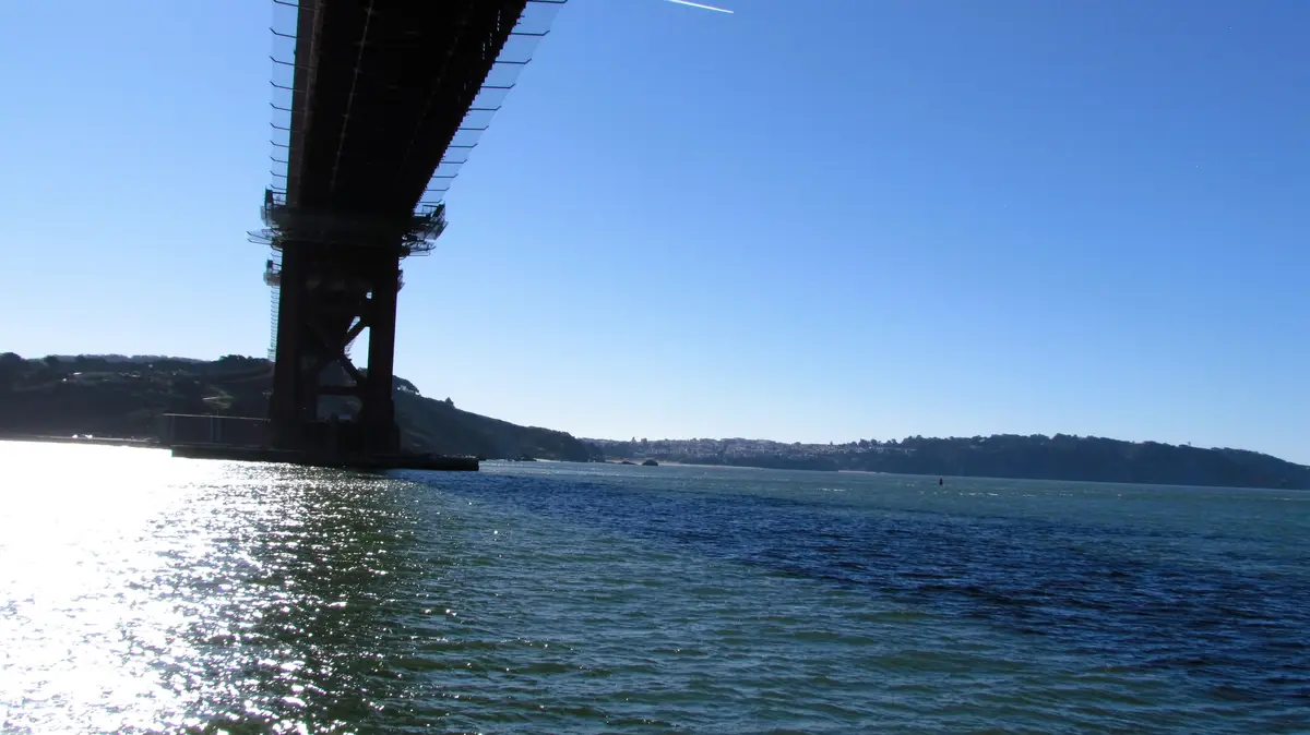 A low-angle photo shows the massive, dark silhouette of a suspension bridge's support tower rising from the water under a…