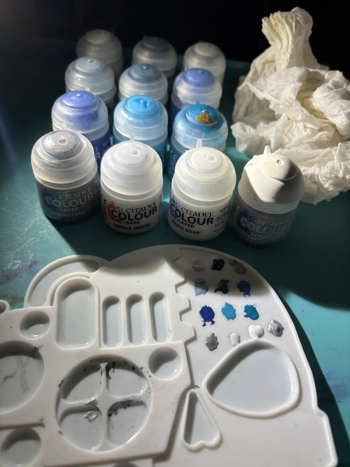A close-up photo shows a white plastic paint palette with various blue, gray, and white paint swatches on it, surrounded by…