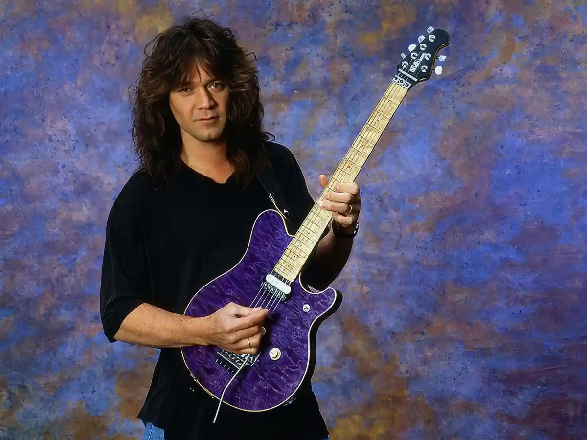 A man with long brown hair wearing a black shirt holds a purple electric guitar against a textured blue and brown…