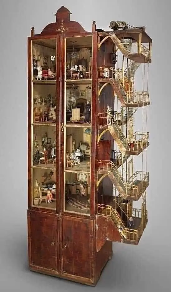 a tall, ornate wooden dollhouse with glass-fronted compartments showing miniature furnished rooms, contrasted by an…