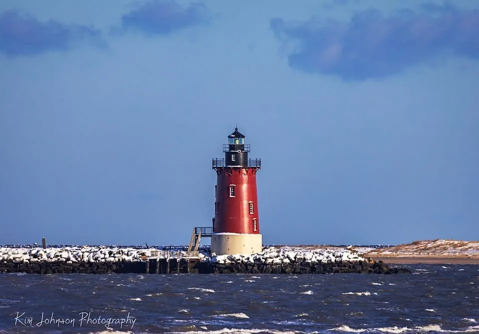 A red and white lighthouse stands on a snow-dusted rocky jetty overlooking choppy water under a blue sky with scattered…
