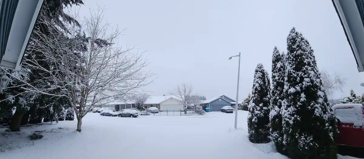 A wide-angle photo shows a snowy suburban neighborhood with houses, snow-covered cars, and evergreen trees under an overcast…