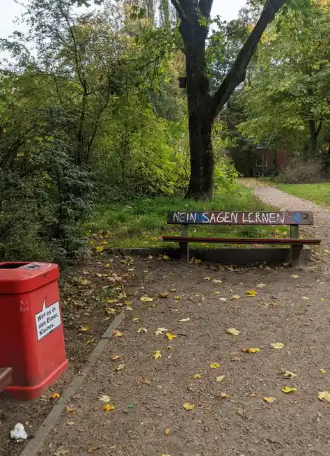 A wooden park bench with graffiti reading 'NEIN SAGEN LERNEN' sits beside a red trash can labeled 'Wurf es in den Eimer…