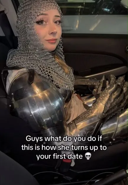 A woman wearing full medieval-style armor, including chainmail hood and metal gauntlets, sits in the passenger seat of a…