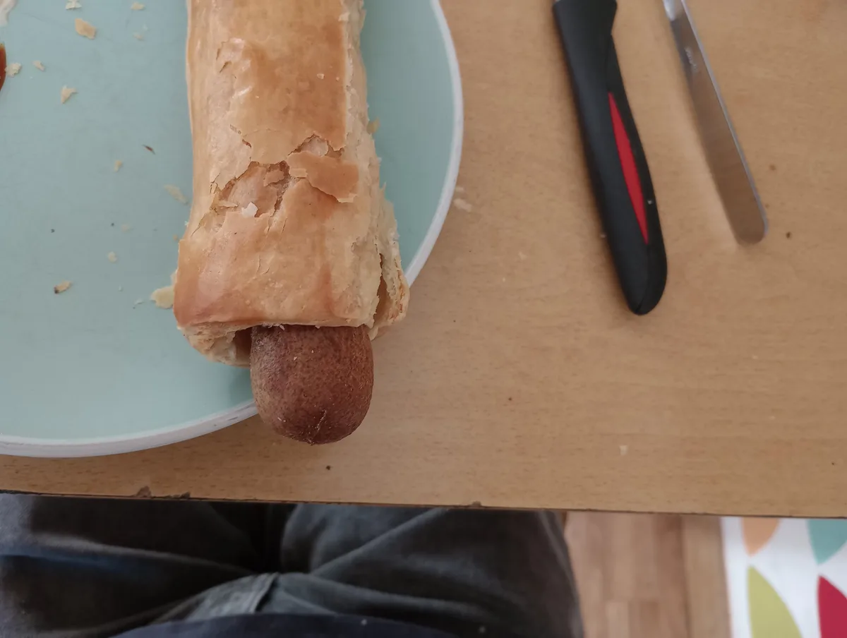 A partially eaten sausage in a pastry wrapper sits on a light blue plate, with a knife and fork nearby on a wooden table…