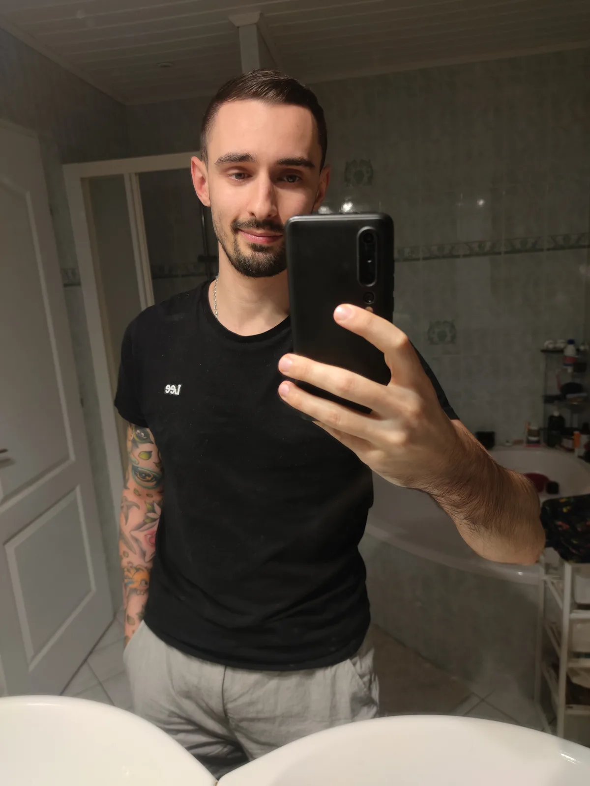 A man with a beard and tattoos on his left arm takes a mirror selfie in a bathroom, holding a black smartphone. He wears a…
