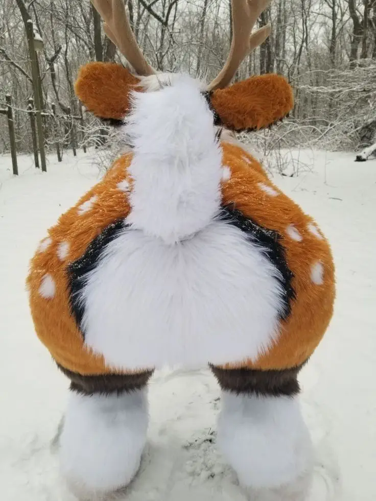 A person in a full-body deer costume stands in a snowy forest, viewed from behind, showcasing a large, fluffy white butt…