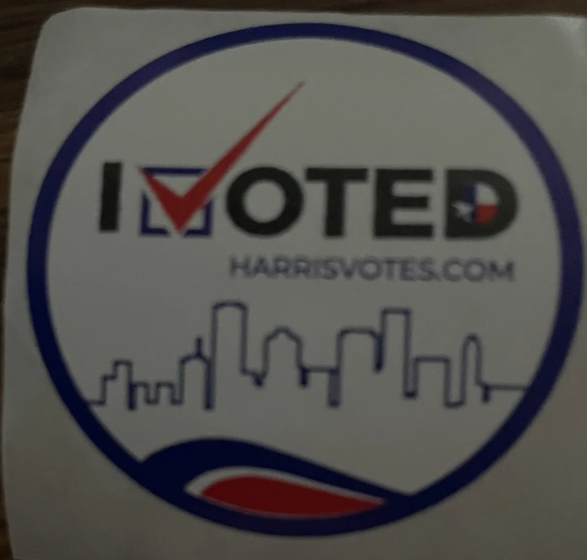 A close-up of a white circular sticker with a blue border, featuring the text 'I VOTED' where the 'V' is replaced by a red…