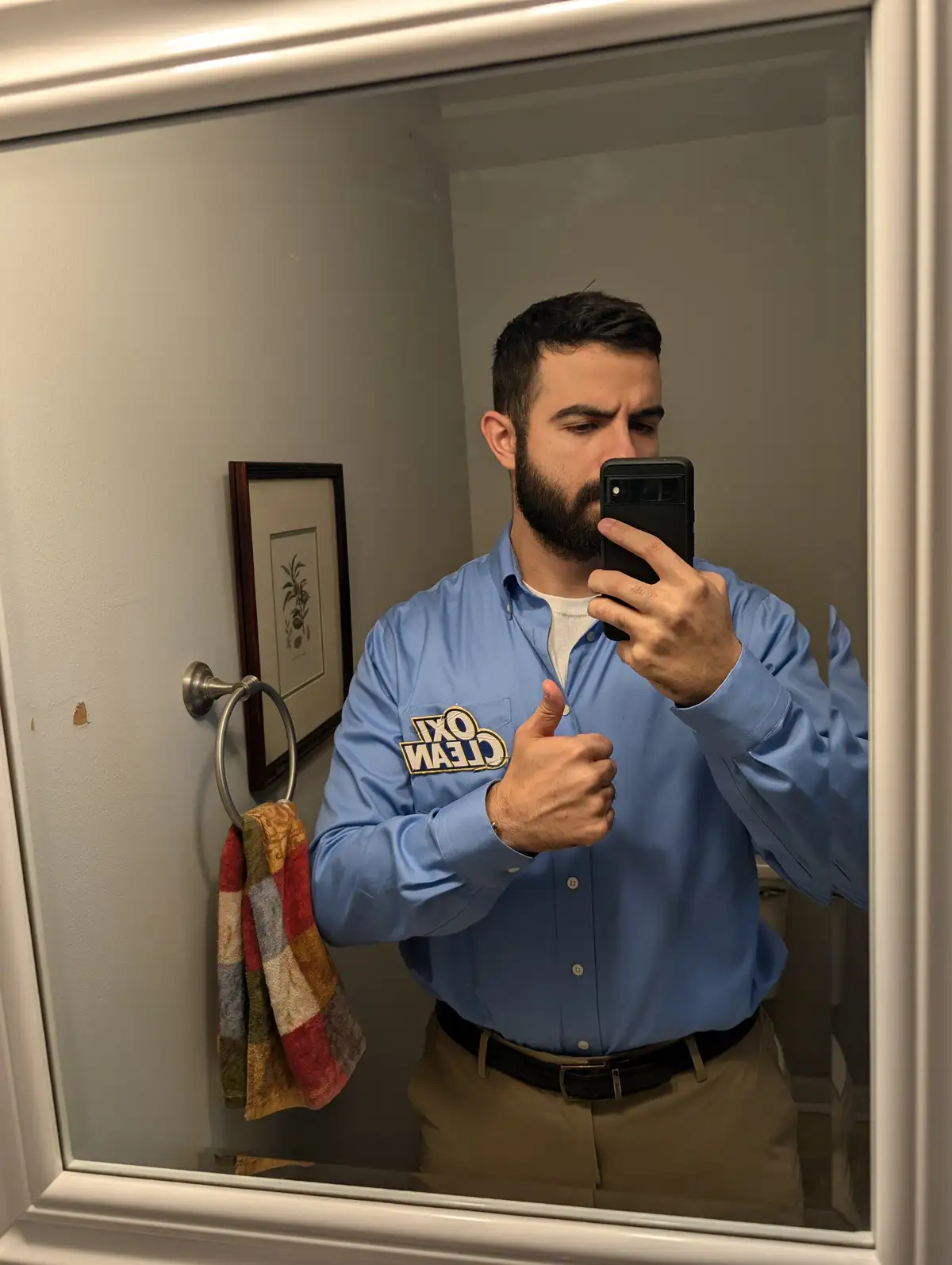 A man in a blue shirt with a 'clean ox' logo on the pocket takes a mirror selfie in a bathroom, giving a thumbs-up while…
