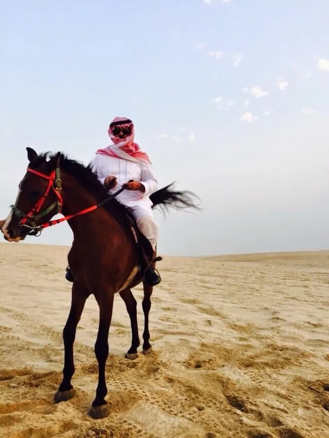 A person wearing traditional white attire and a red-and-white checkered headscarf rides a dark brown horse across a vast…
