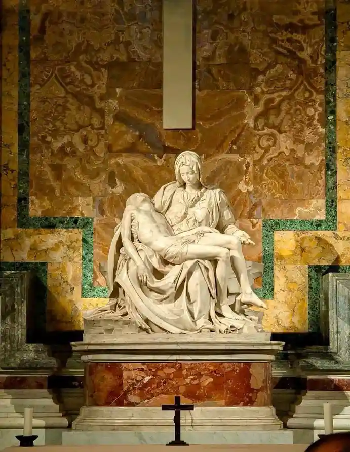 A marble sculpture depicts the Virgin Mary cradling the lifeless body of Jesus Christ, set against a backdrop of ornate…