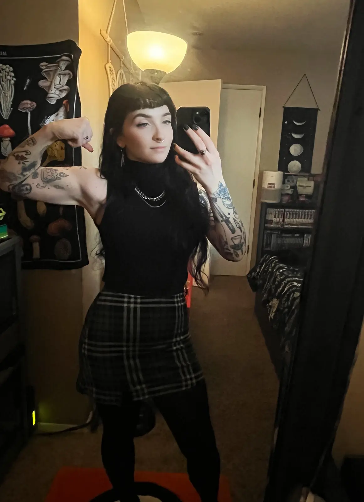 A person with long dark hair and tattoos on both arms takes a mirror selfie in a dimly lit room, flexing their bicep while…