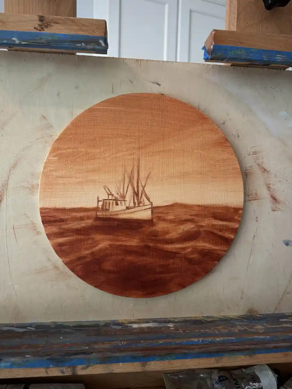 A close-up of a circular wood-burned artwork depicting a fishing boat on the water, mounted on a wooden board with blue…