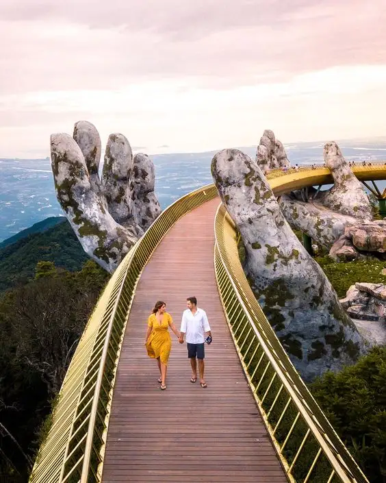 A couple walks hand-in-hand along a golden, elevated wooden bridge that arches between two giant stone hands, with a vast…