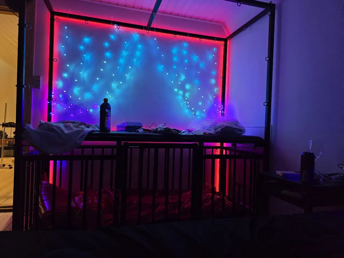 A dimly lit bedroom features a black metal bed frame with a mattress and bedding. Behind the bed, a wall is illuminated with…