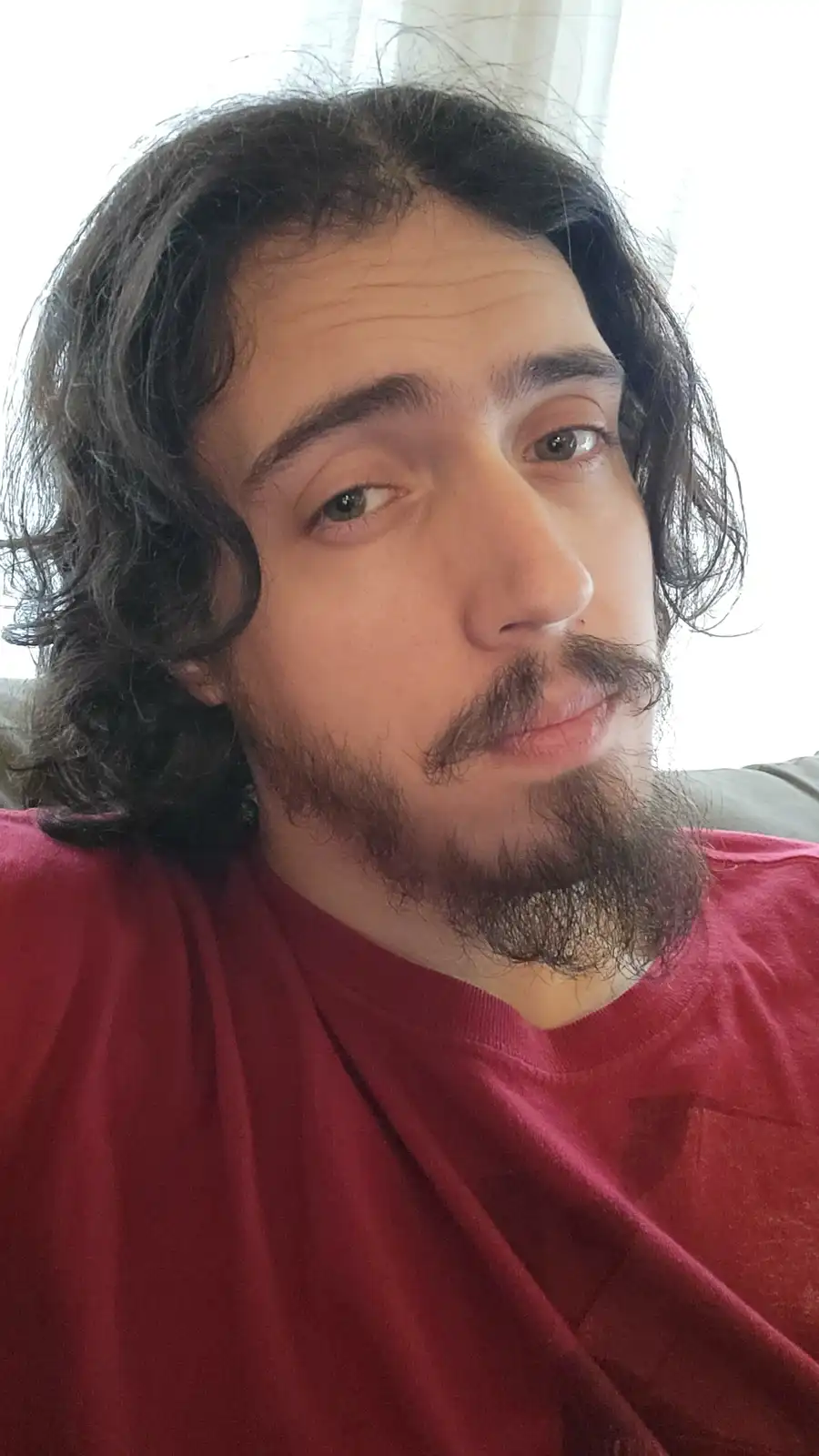 A selfie of a man with long dark hair, a beard, and mustache, wearing a red t-shirt, looking directly at the camera with a…