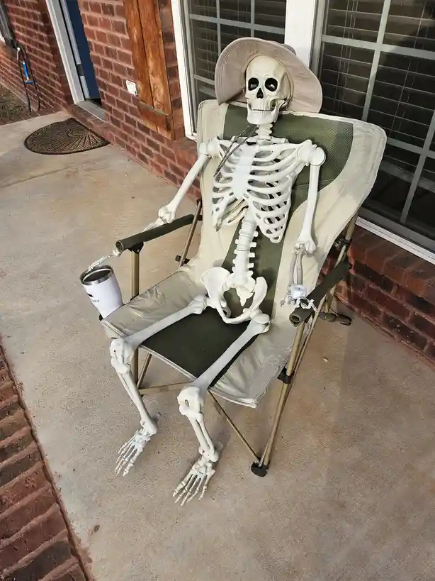 A plastic skeleton wearing a beige sun hat sits relaxed in a folding camping chair on a concrete porch, holding a white…