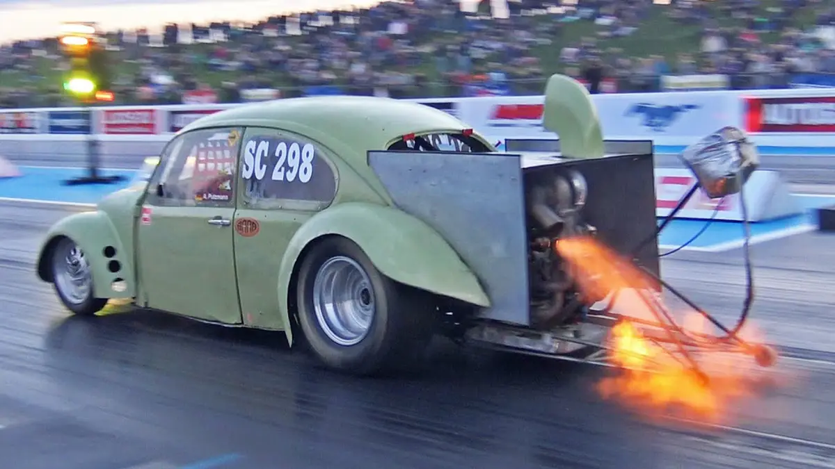 A heavily modified, olive-green Volkswagen Beetle race car with the number SC 298 on its side is speeding down a drag strip…