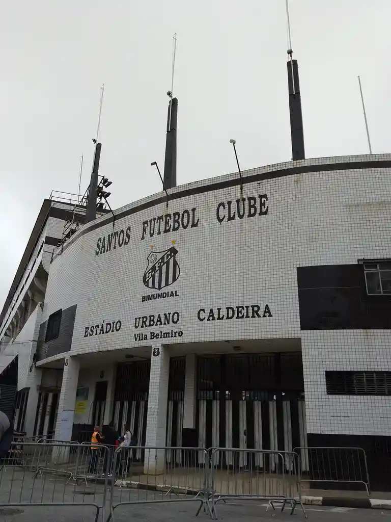 The image shows the exterior of Vila Belmiro stadium, home to Santos Futebol Clube, with its white tiled facade and black…