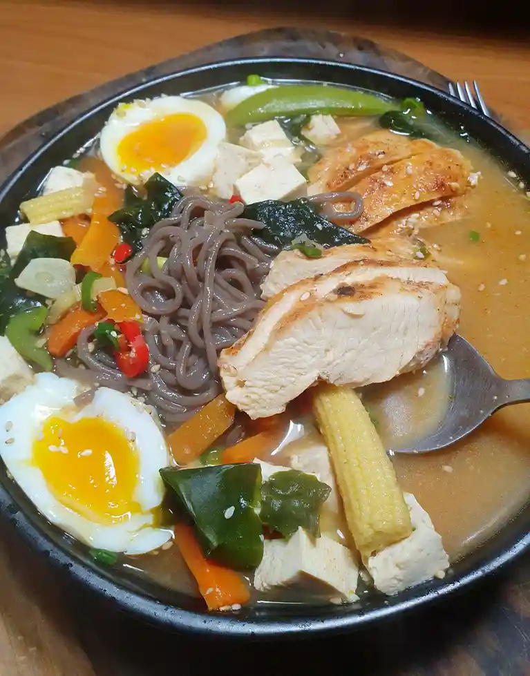 A close-up photo of a steaming bowl of Korean-style noodle soup with dark soba noodles, sliced chicken, tofu, baby corn…
