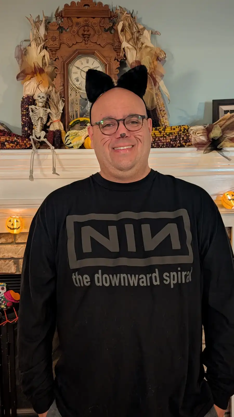 A man wearing black cat ears and cat whiskers smiles while dressed in a black long-sleeved shirt that reads 'NIN the…