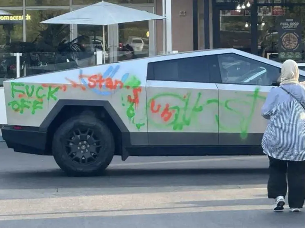 A silver Tesla Cybertruck is covered in colorful graffiti, including the words 'FUCK OFF' in green spray paint on its rear…