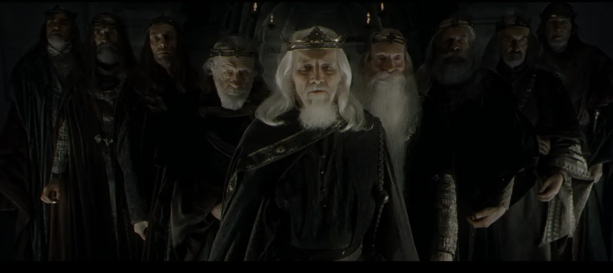 A still from a fantasy film shows a stern, elderly king with a crown and long white beard, flanked by several similarly…