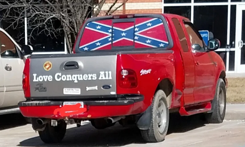 The rear view of a red Ford pickup truck with a gray tailgate displaying the phrase 'Love Conquers All' in white text. The…