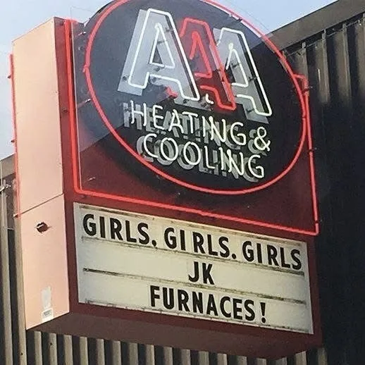 A neon sign for 'AA Heating & Cooling' features a marquee below that reads 'GIRLS, GIRLS, GIRLS JK FURNACES!'. The sign is…