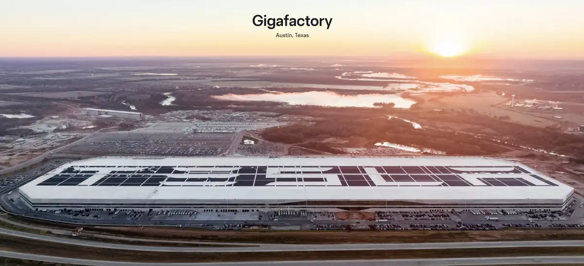 An aerial photo shows a massive, modern industrial building with a white roof featuring a large, stylized logo made of dark…