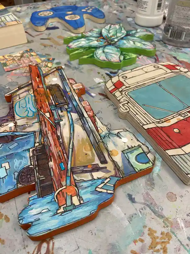 A close-up photo shows several hand-painted wooden coasters on a paint-splattered artist's table. The most prominent coaster…