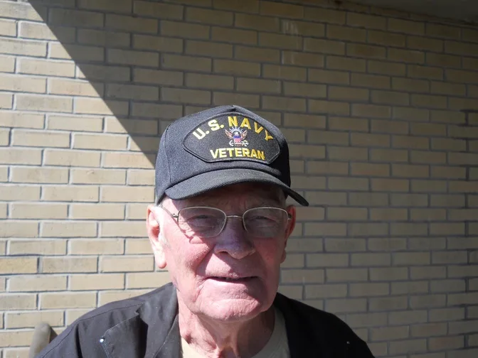 An elderly man with glasses and a weathered face wears a dark blue U.S. Navy Veteran cap with yellow lettering and an…
