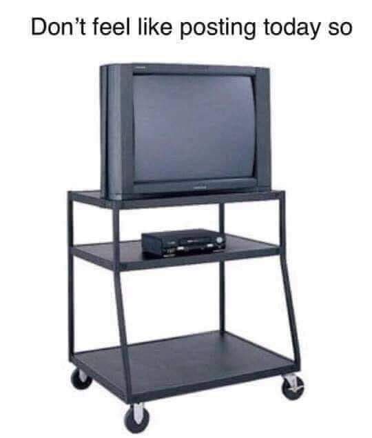 An old, bulky CRT television sits on a black, three-tiered rolling cart with wheels. The middle shelf holds a small black…