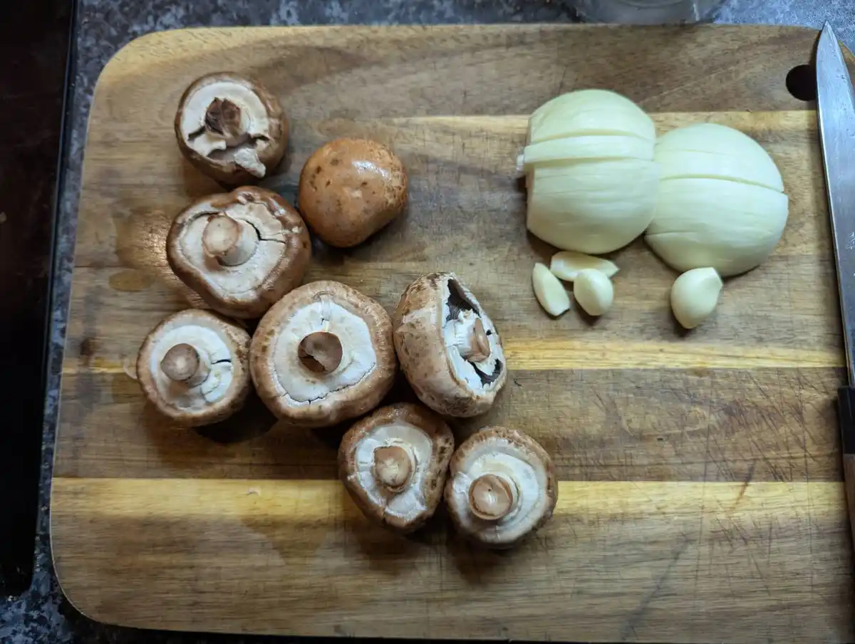 A wooden cutting board holds several sliced and whole brown mushrooms, a halved onion, and garlic cloves, with a knife…