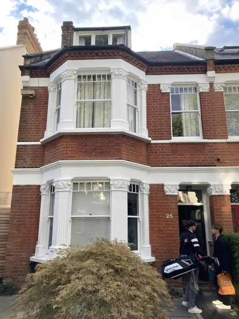 A red brick Victorian-style house with white trim and bay windows, number 25, has two men exiting its front door. One man…