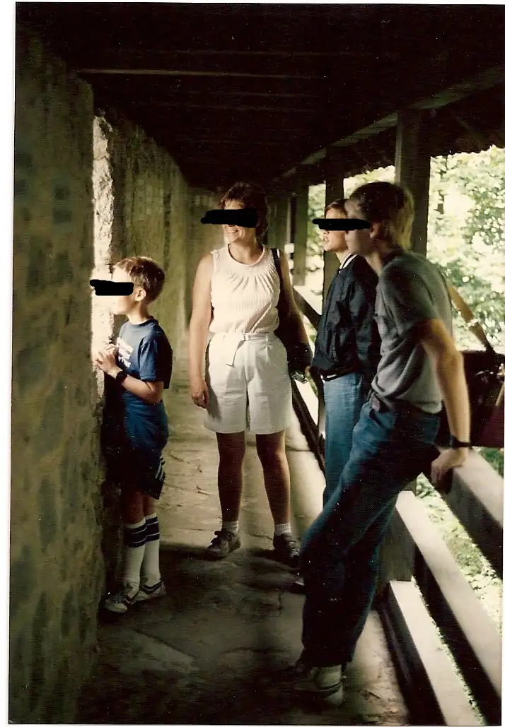 A grainy, vintage family photo shows four people standing in a covered outdoor walkway. The boy on the left is pressed…