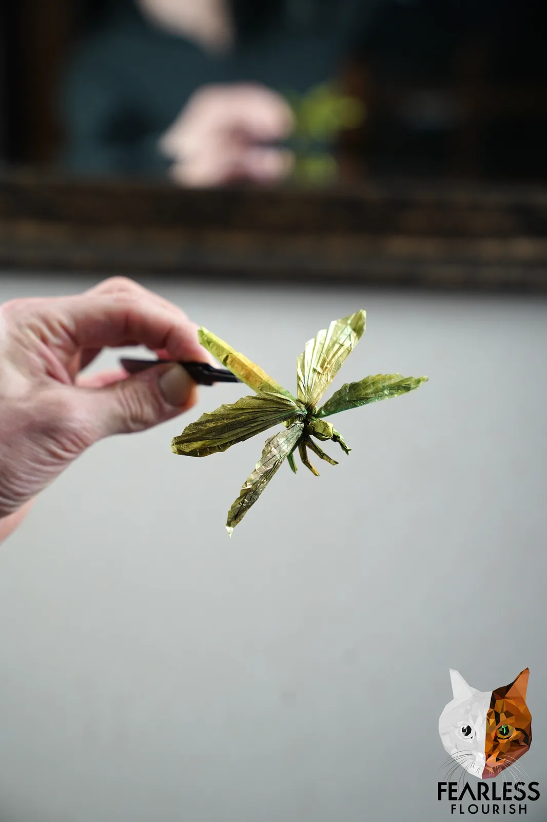 A hand holds a delicate, green origami dragonfly made from folded paper, suspended in front of a blurred background where…