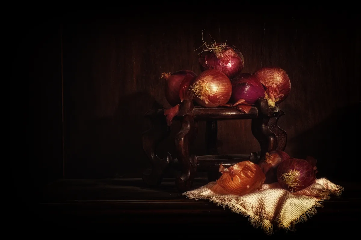 A dramatic, chiaroscuro-style still life photograph shows several red onions arranged on a dark wooden stool and a burlap…