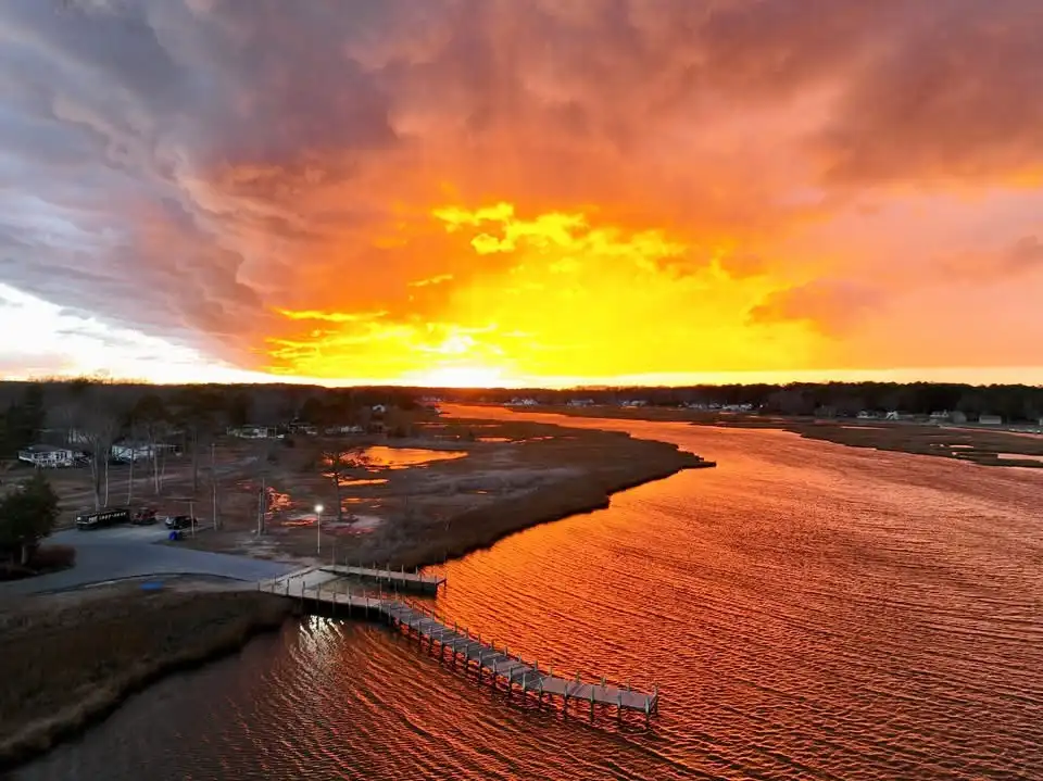 Aerial view of a wooden pier extending into a body of water during a vibrant sunset, with fiery orange and yellow clouds…