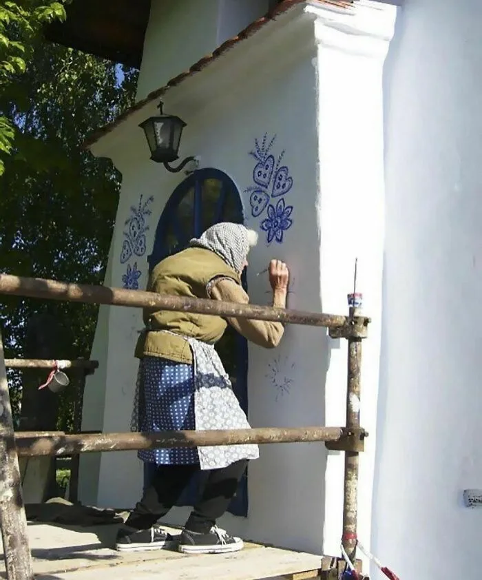 An elderly woman wearing a headscarf and apron stands on a wooden scaffold, painting intricate blue floral and heart…