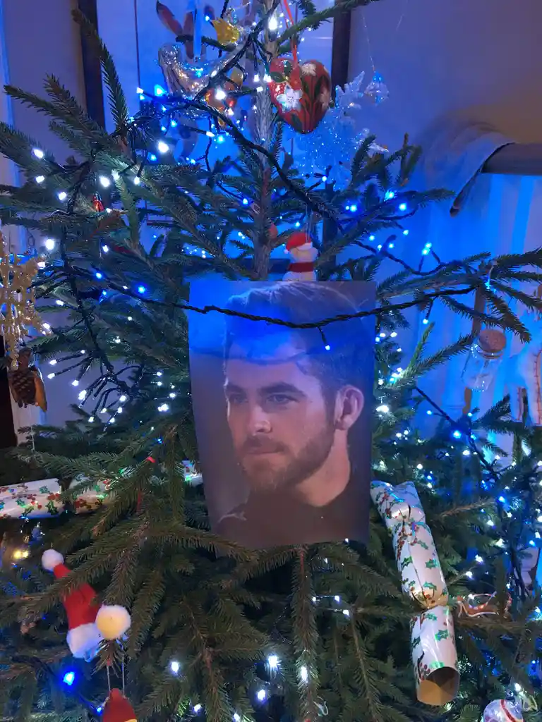 A decorated christmas tree with blue lights and ornaments has a printed photo of a man with a serious expression taped to…